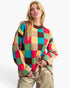 A woman modeling a colorful oversized knit sweater with a large checkered pattern in red, green, yellow, brown, and beige, paired with metallic gold pants against a plain white background.