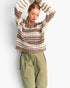 A woman wearing a brown and cream striped crochet knit sweater and olive green cargo pants, posing with her arms raised against a light gray background.