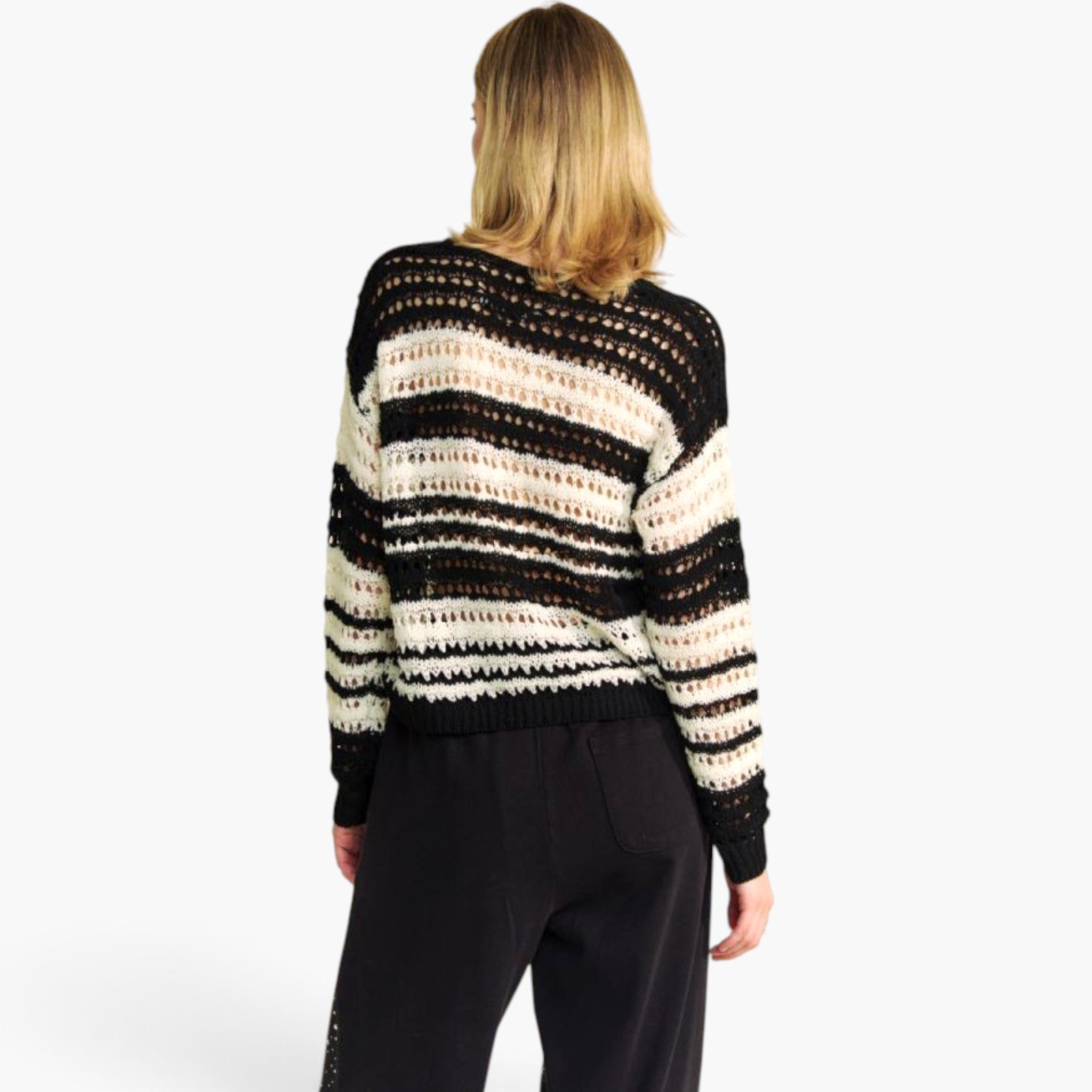 A woman viewed from behind wearing a black and cream striped knit sweater with a textured open-knit pattern, paired with black pants, against a plain white background.