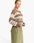 A woman wearing a brown and cream striped crochet knit sweater and olive green pants, photographed against a plain white studio background.