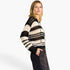 A woman wearing a black and cream striped knit cropped sweater with a drawstring hood, paired with black pants, photographed against a clean white studio background.