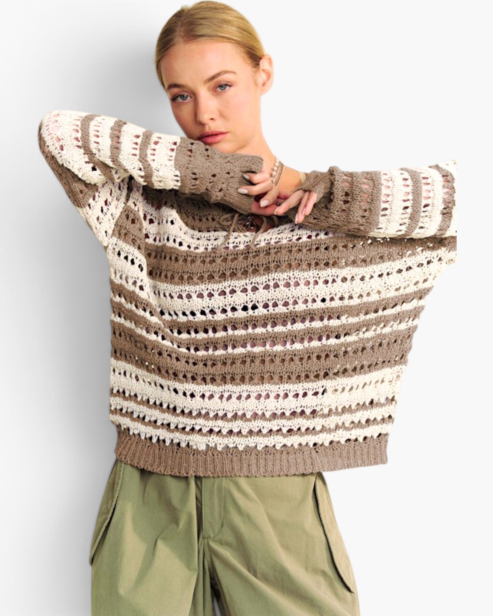 A woman wearing a chunky knit striped sweater in taupe and cream, paired with olive green cargo pants, against a light gray studio background.