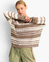 A woman wearing a chunky knit striped sweater in taupe and cream, paired with olive green cargo pants, against a light gray studio background.