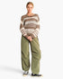 A model stands against a light gray studio background wearing a chunky knit crewneck sweater with horizontal brown, beige, and cream stripes, paired with wide-leg olive green pants and black canvas sneakers.