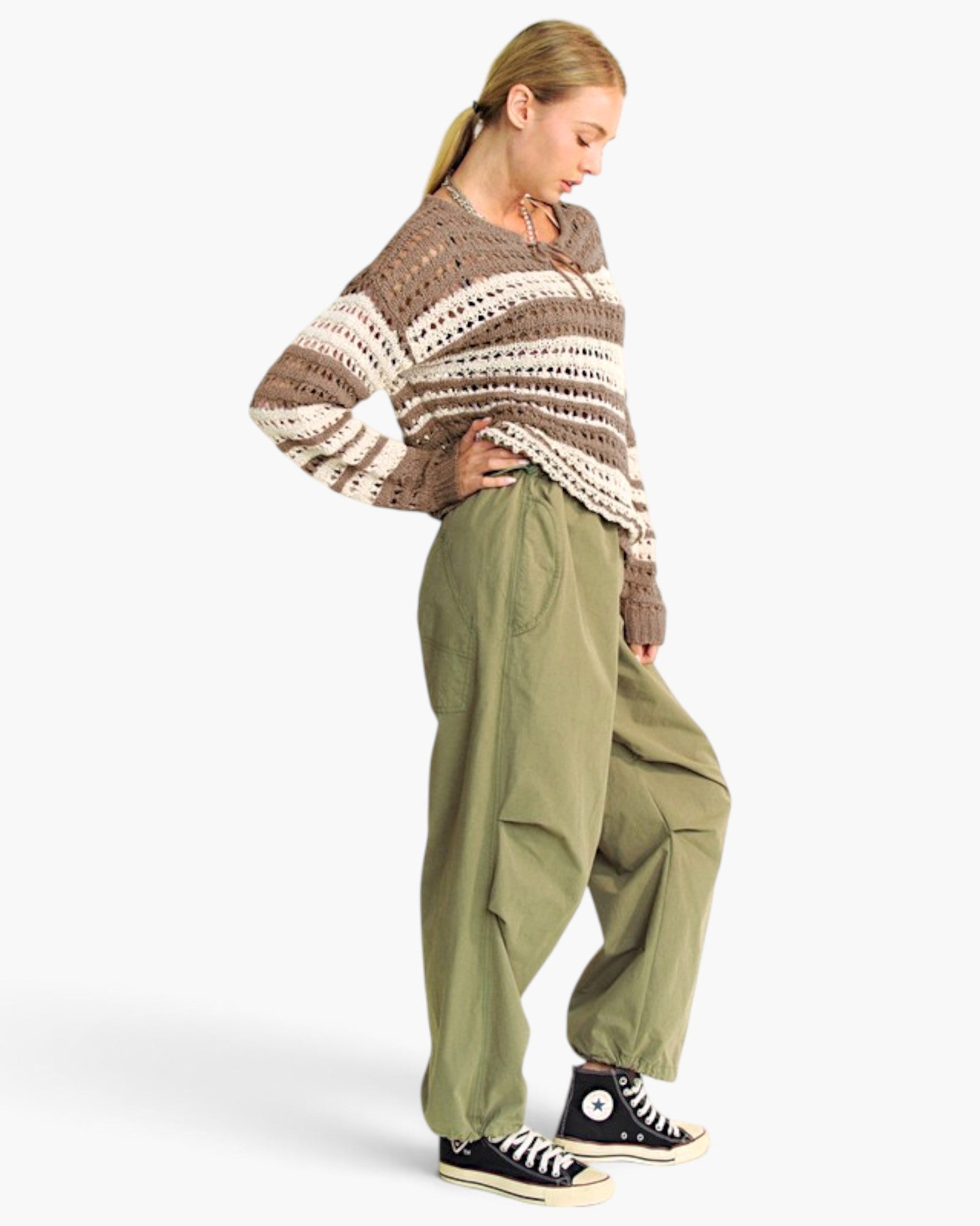 A woman modeling a brown and cream striped crochet knit sweater paired with olive green baggy pants and black high-top sneakers, set against a plain light gray studio background.