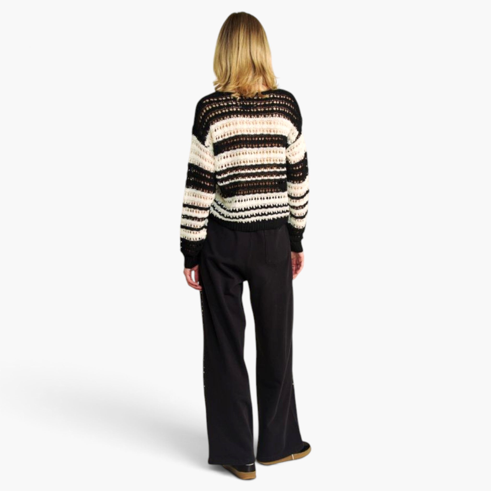 A woman viewed from behind wearing a black and cream striped knit sweater and black wide-leg trousers, standing against a plain light gray studio background.