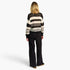 A woman viewed from behind wearing a black and cream striped knit sweater and black wide-leg trousers, standing against a plain light gray studio background.