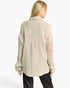 A woman shown from the back wearing a light beige textured button-up shirt with sheer eyelet knit sleeves, paired with black pants, against a plain white studio background.