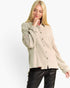A blonde woman wearing a beige textured button-up shirt with openwork knit sleeves and black pants, posing against a plain white studio background.