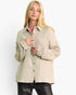A blonde woman modeling a light beige textured button-up shirt with knit cuffed sleeves, worn over a pink top and black pants, against a plain white studio background.