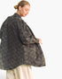 A woman wearing a dark gray washed denim jacket with a black diamond grid pattern, paired with cream-colored pants, shown from a side-back angle against a plain white background.