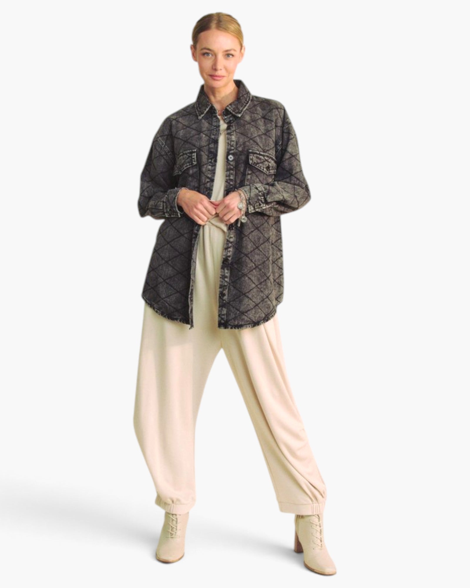 A model stands against a light gray studio background wearing a dark gray quilted button-up shirt jacket over a cream top and cream wide-leg pants with beige lace-up ankle boots.