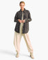 A model stands against a light gray studio background wearing a dark gray quilted button-up shirt jacket over a cream top and cream wide-leg pants with beige lace-up ankle boots.