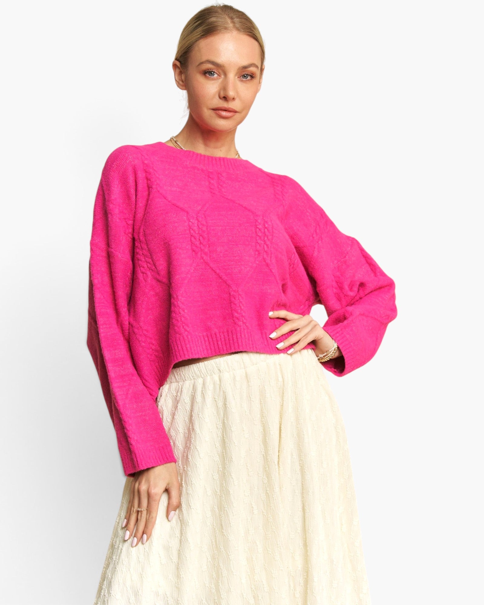 A woman modeling a bright pink cable-knit cropped sweater with balloon sleeves, paired with a cream textured skirt, against a plain white studio background.