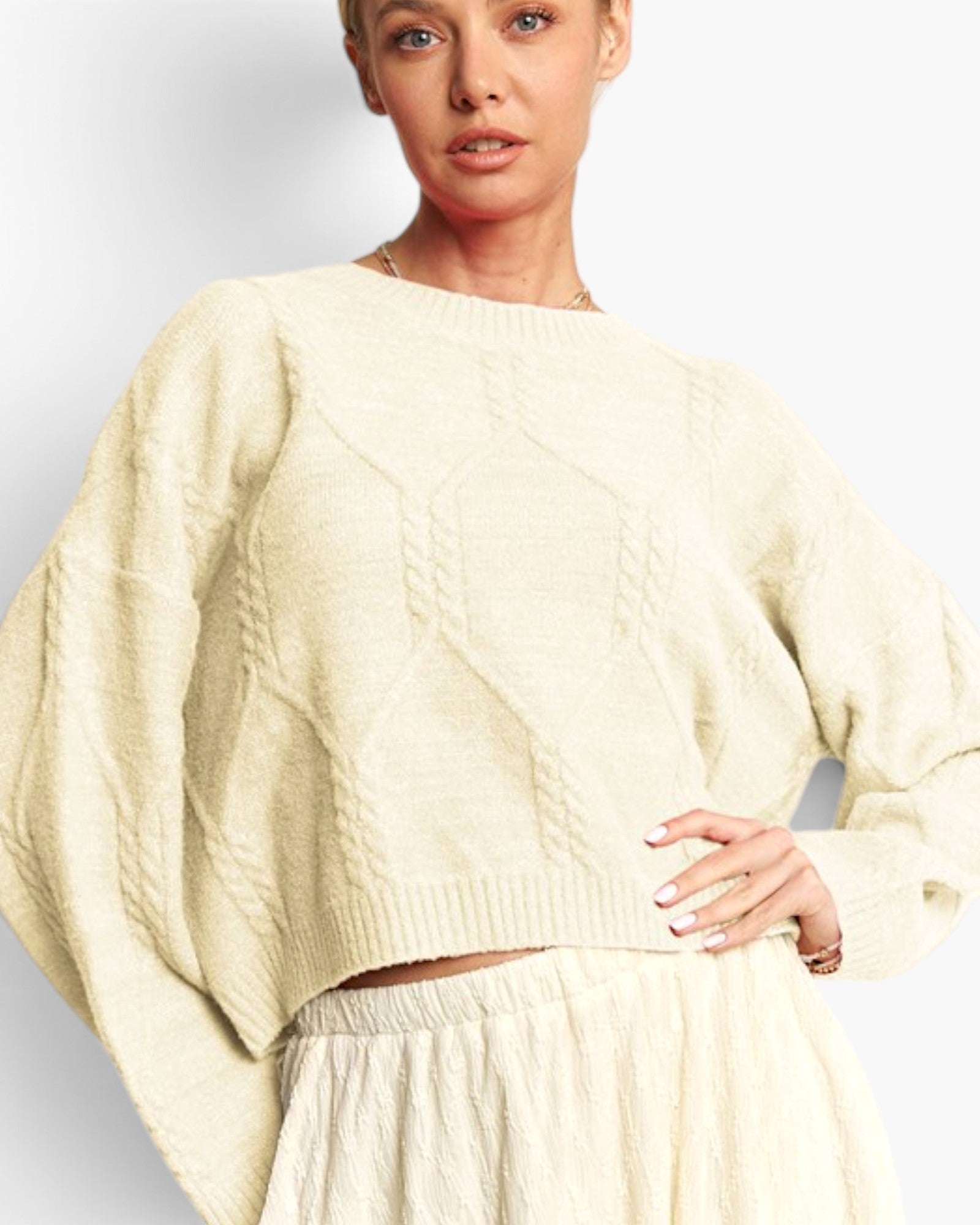 A woman wearing a cream-colored cable-knit cropped sweater with oversized balloon sleeves, paired with a matching textured skirt, against a plain light gray studio background.