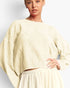 A woman wearing a cream-colored cable-knit cropped sweater with oversized balloon sleeves, paired with a matching textured skirt, against a plain light gray studio background.
