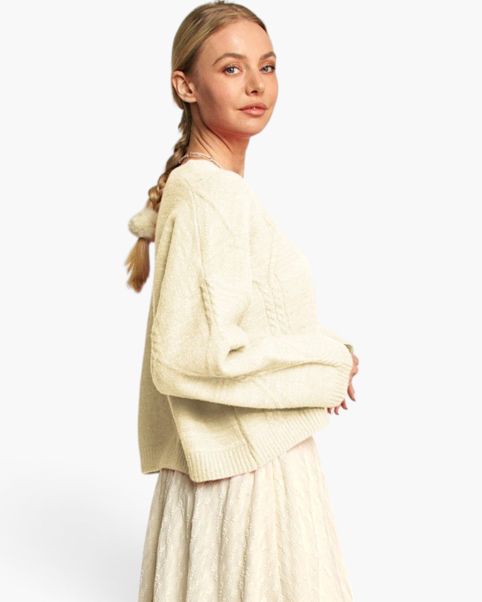A woman wearing a cream cable-knit cropped sweater and matching textured skirt, photographed against a plain light gray studio background.