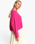 A woman wearing a bright pink cable-knit cropped sweater and a cream-colored skirt, photographed against a clean white studio background.