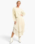A woman modeling a cream-colored two-piece knit set consisting of a cropped long-sleeve sweater and a matching textured midi skirt with an asymmetrical layered hem, paired with white ankle boots, against a plain white studio background.