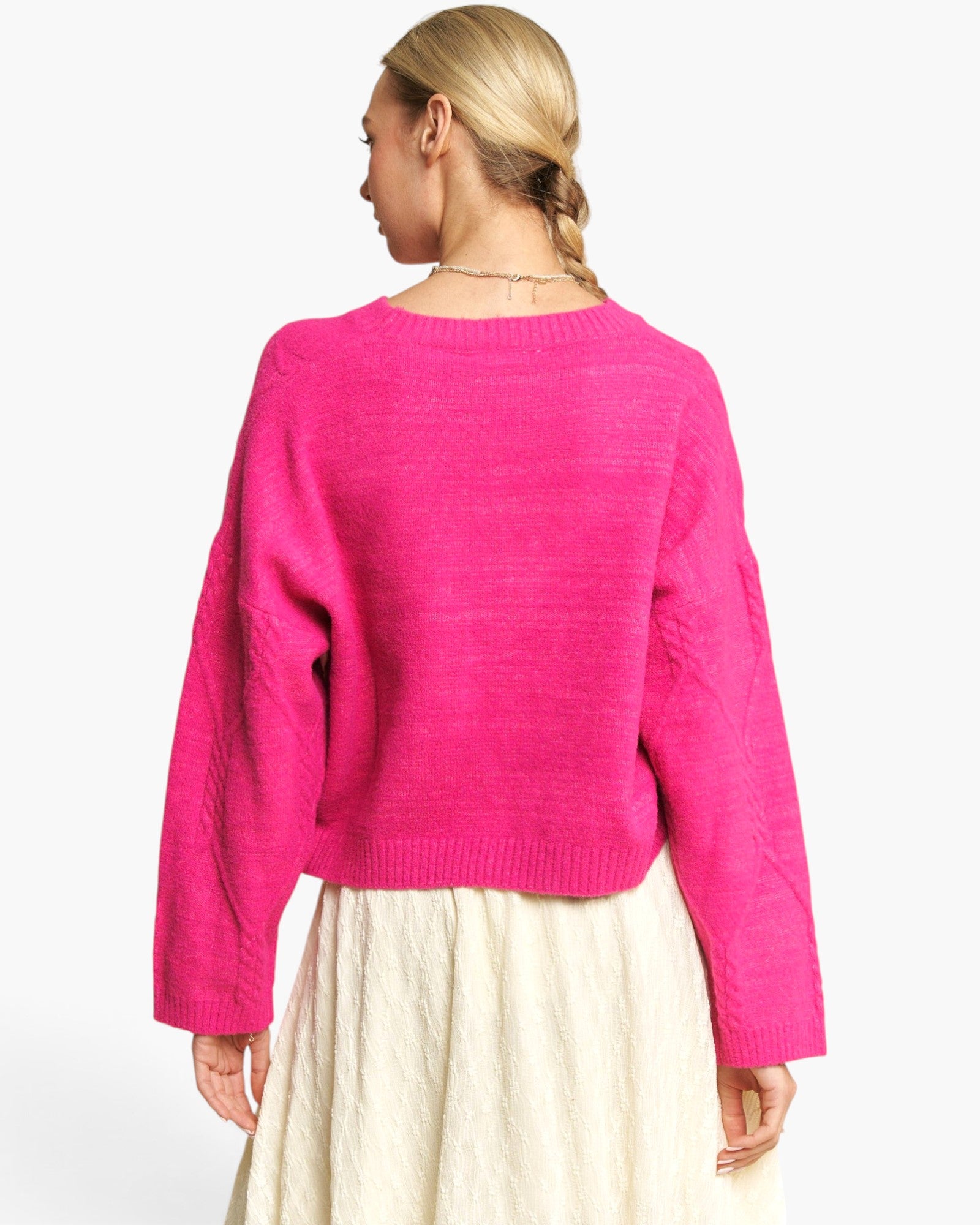 A woman shown from the back wearing a bright pink knit sweater and a cream-colored skirt, photographed against a clean white studio background.