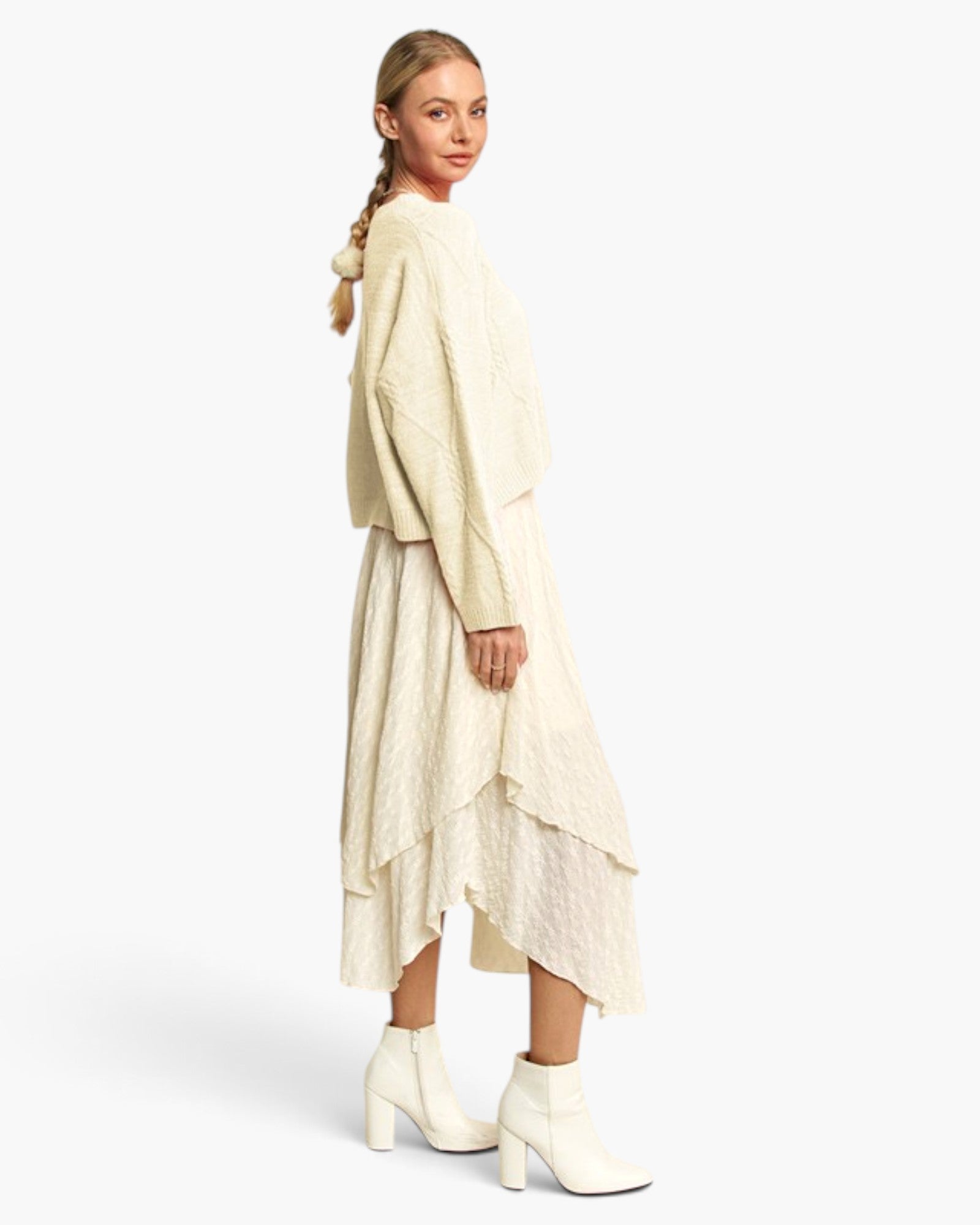 A model is shown from the back wearing a cream-colored textured knit sweater and matching layered asymmetrical skirt, paired with white ankle boots, against a plain light gray studio background.
