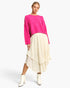 A model wearing a bright pink textured knit sweater and a cream-colored tiered midi skirt, paired with white ankle boots, against a plain white studio background.