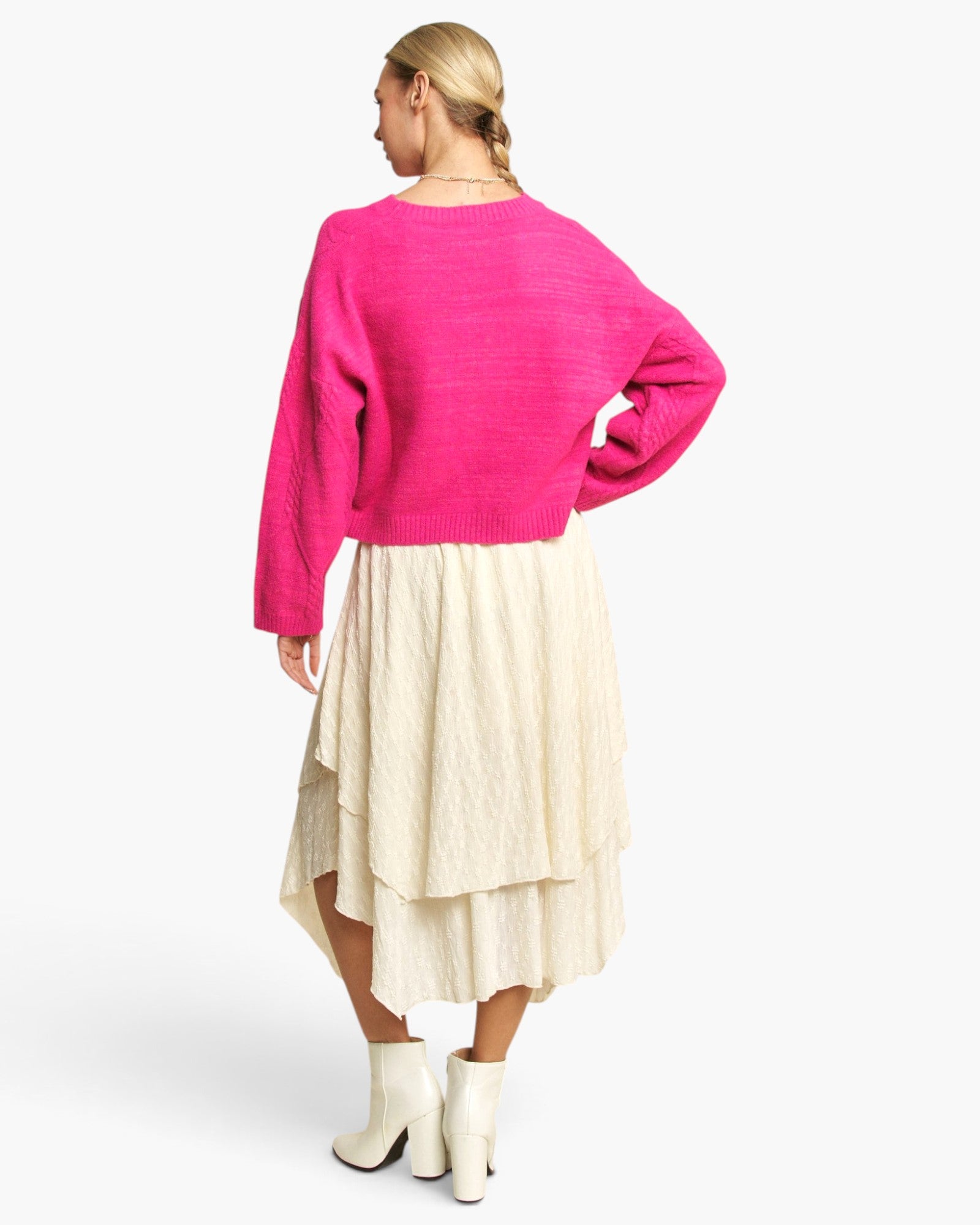 A woman shown from the back wearing a bright pink cropped knit sweater and a cream-colored layered skirt, paired with white ankle boots, against a light gray studio background.