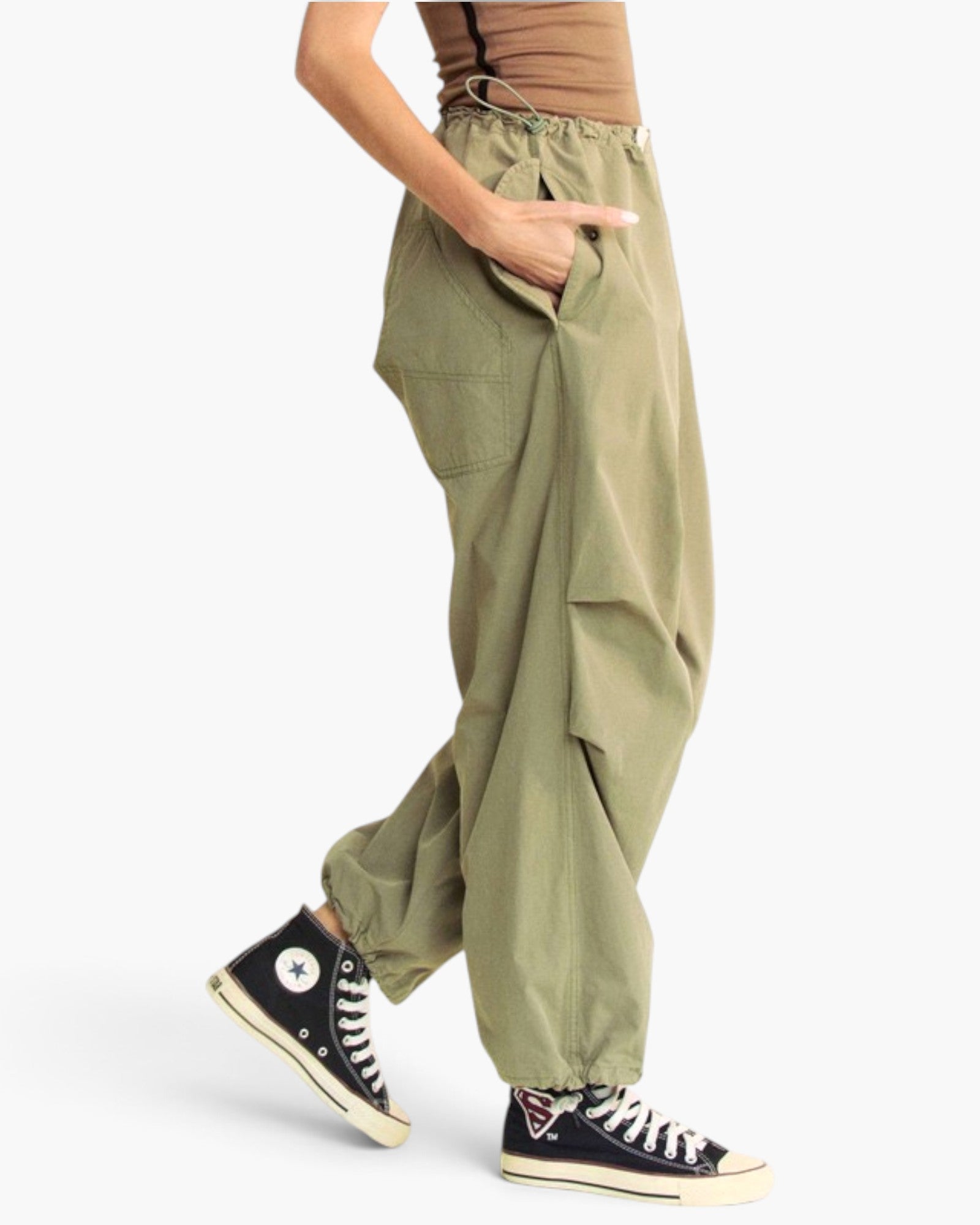 A person wearing loose, baggy olive green cargo pants with side pockets and elastic cuffs, paired with black high-top sneakers, shown from the waist down on a light gray studio background.