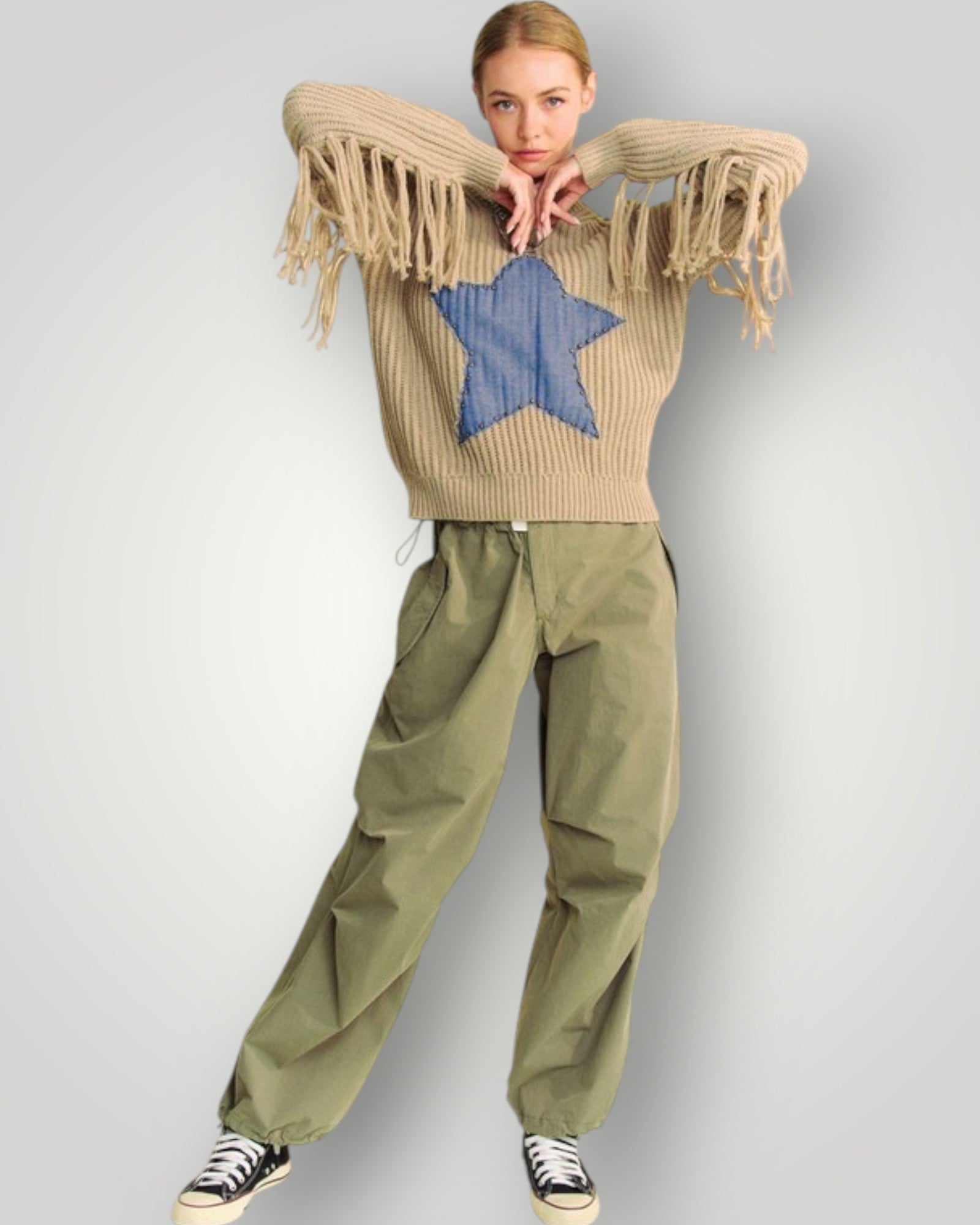 Person wearing a beige sweater with fringe and a blue star, paired with green pants on a light gray background