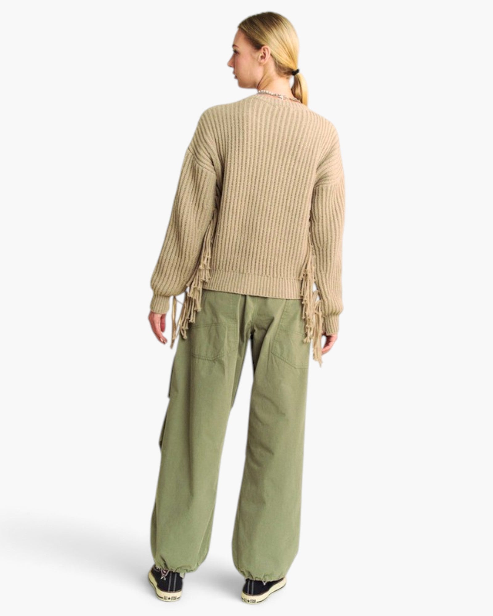 A woman shown from the back wearing a beige ribbed knit sweater with fringe details and olive green wide-leg cargo pants, photographed against a clean white studio background.