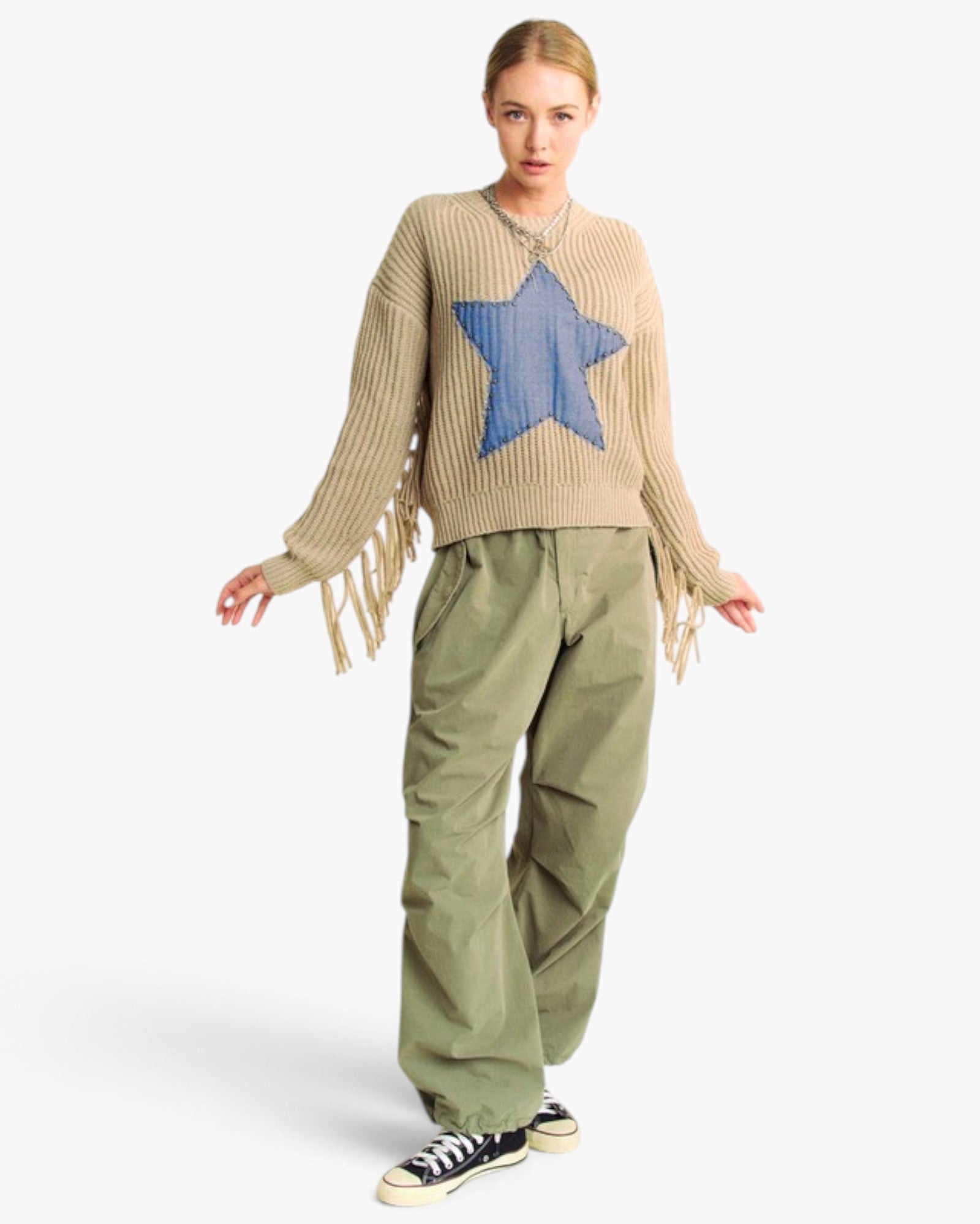 A model wearing a beige ribbed knit sweater with a large blue star patch on the chest and fringe details on the sleeves, paired with olive green cargo pants and black canvas sneakers, against a light gray background.
