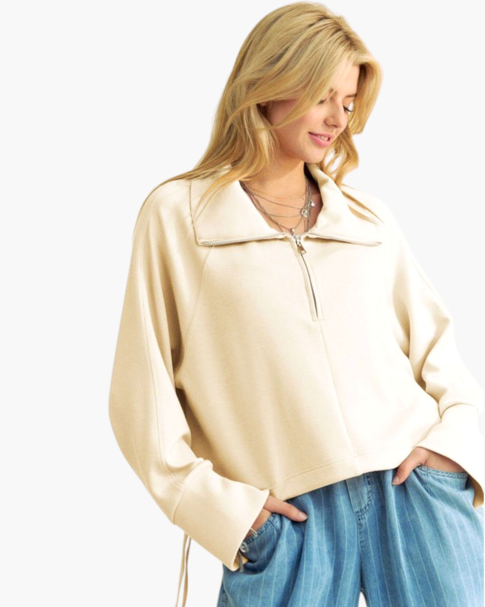 A woman wearing a cream-colored, oversized quarter-zip sweatshirt with a wide collar and blue jeans, photographed against a solid white background.