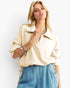 A blonde model wearing a cream-colored, oversized zip-up collared top and blue denim shorts, posed against a clean white studio background.