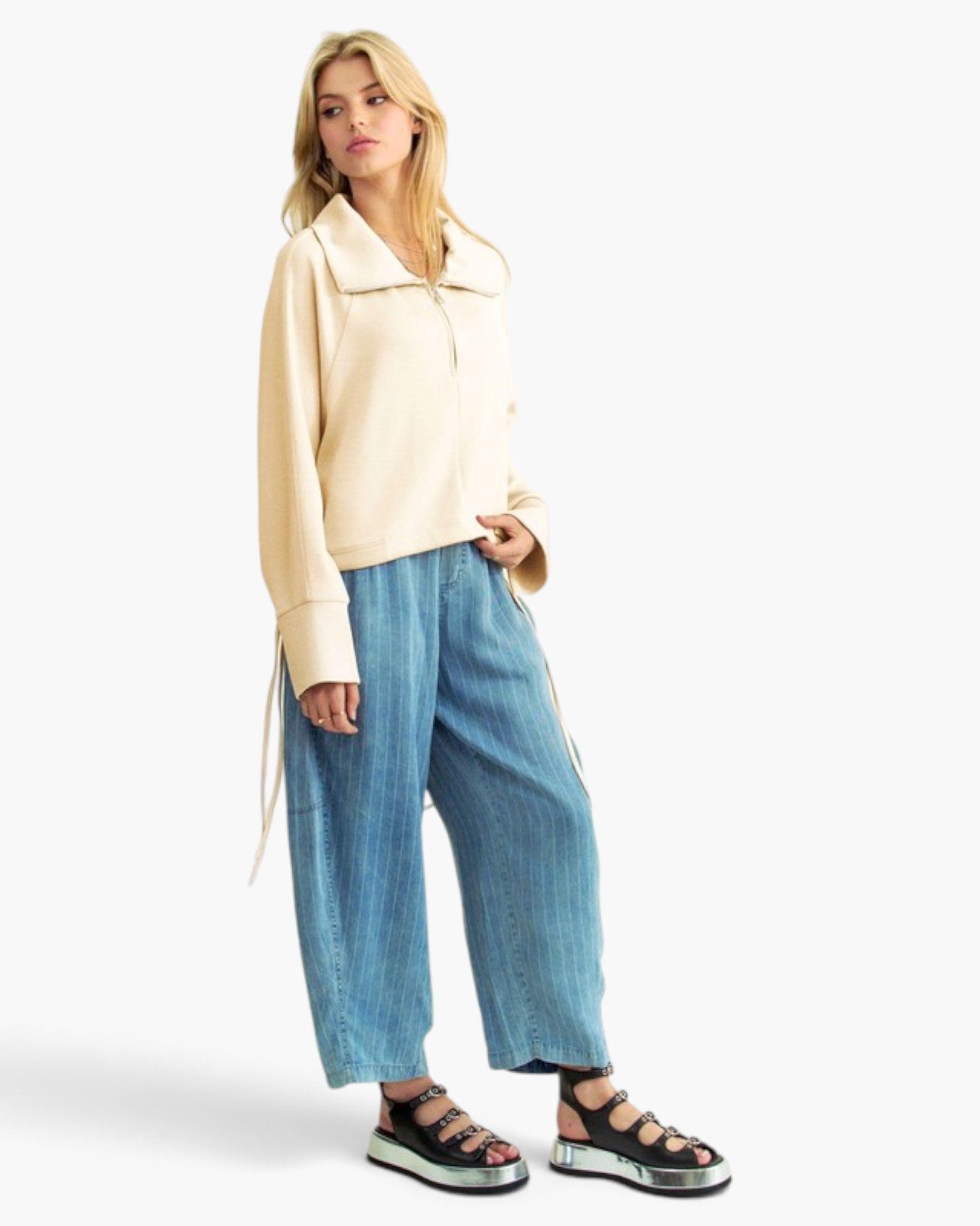 A woman modeling a cream-colored, long-sleeve collared jacket with a drawstring, paired with wide-leg blue jeans and black platform sandals, against a plain white studio background.