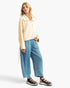 A woman modeling a cream-colored, long-sleeve collared jacket with a drawstring, paired with wide-leg blue jeans and black platform sandals, against a plain white studio background.