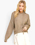 A woman wearing a textured, oversized knit hoodie in a warm taupe-brown color, paired with cream cargo pants, against a plain white studio background.
