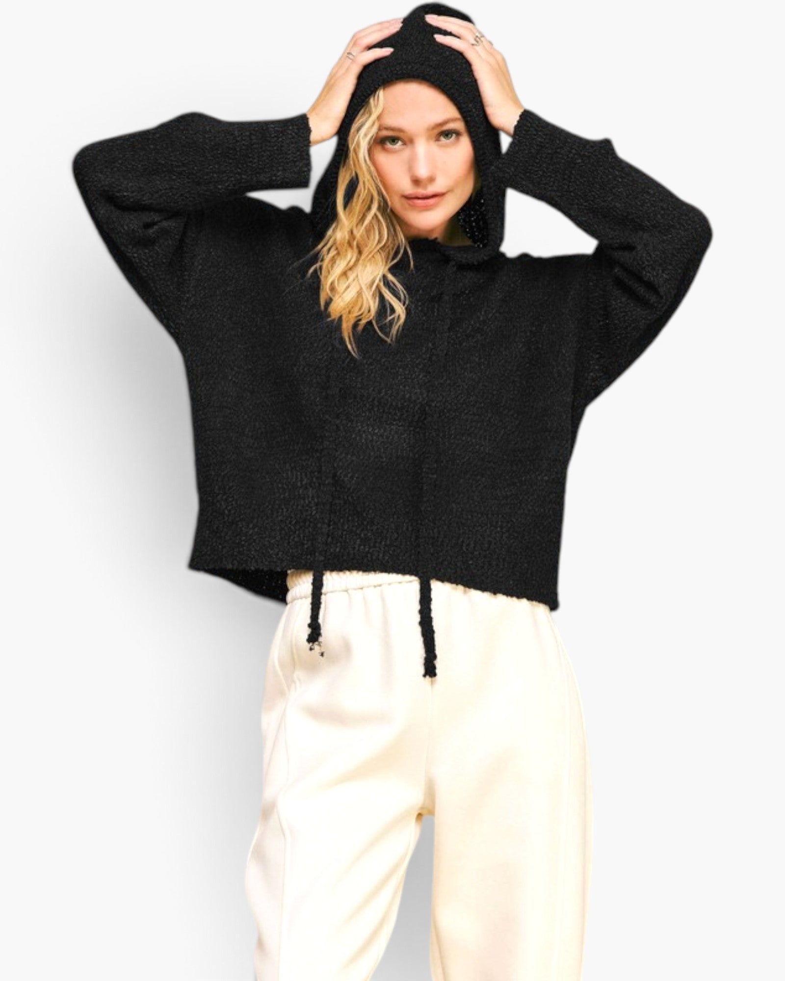 A woman modeling a black cropped knit hoodie with a drawstring, paired with cream sweatpants, against a plain light gray background.