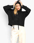 A woman modeling a black cropped knit hoodie with a drawstring, paired with cream sweatpants, against a plain light gray background.