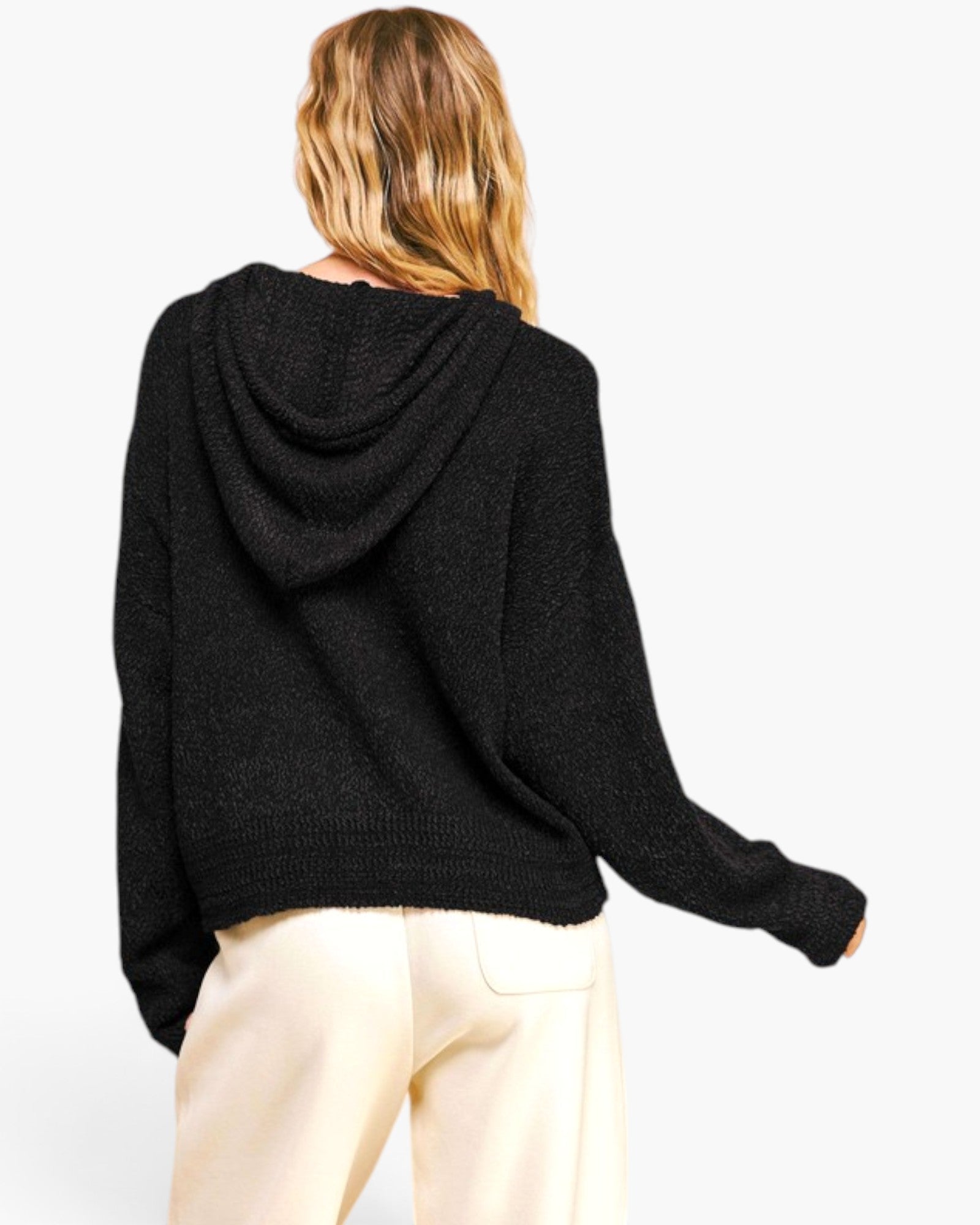 A woman shown from the back wearing a black knit hooded sweater and cream-colored pants against a plain light gray studio background.