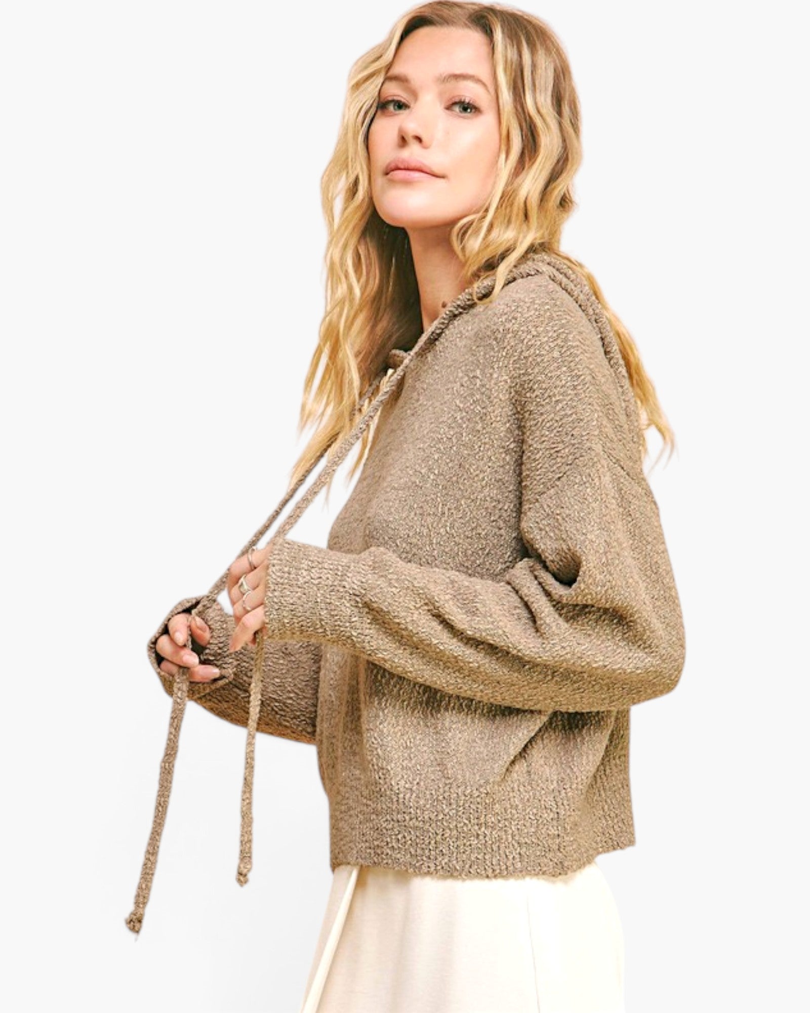 A woman wearing a textured, oversized knit hoodie in a warm taupe-brown color, holding the drawstrings. The background is a clean, solid white.