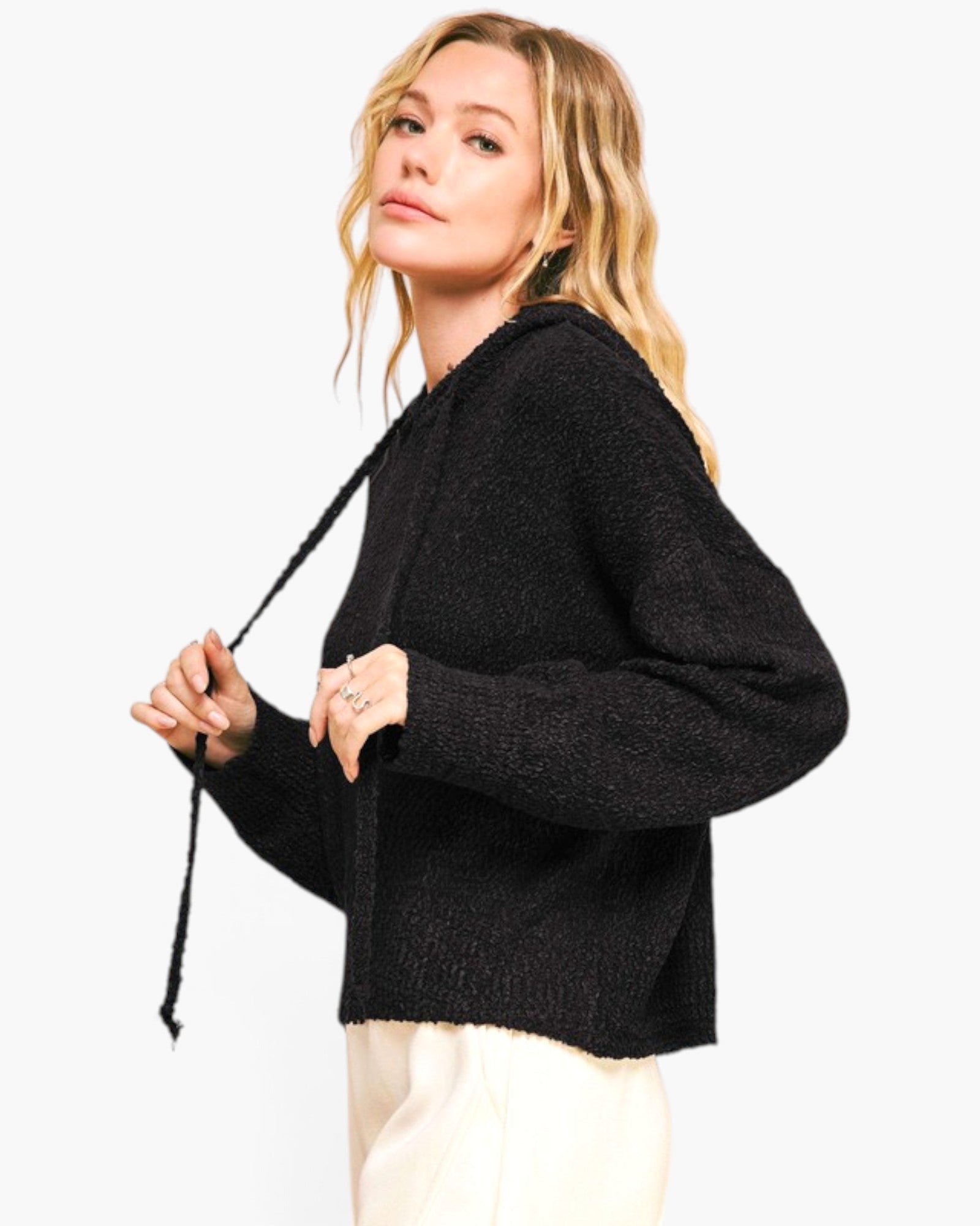 A woman wearing a black textured knit sweater with a drawstring hood, paired with cream pants, photographed against a clean white background.