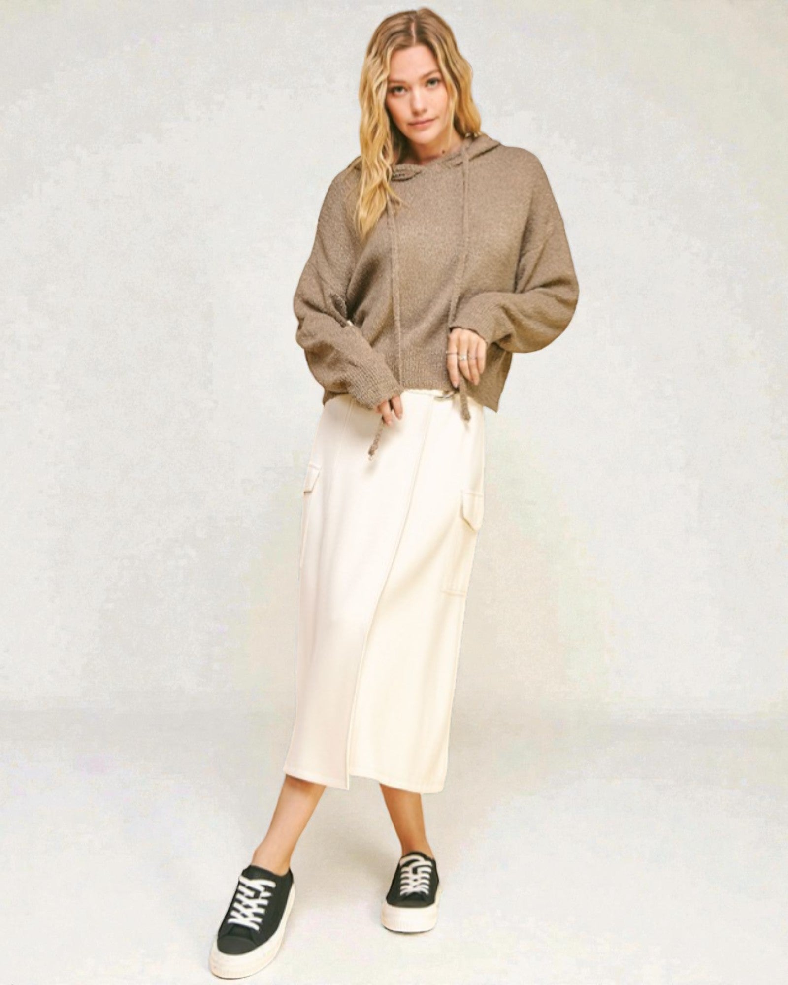 Woman wearing a brown hoodie and white pants against a white background
