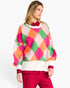 A woman wearing a colorful knit sweater with a pink, orange, green, and white harlequin diamond pattern, paired with red pants, against a plain white background.