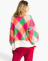 A woman shown from the back wearing a colorful diamond-patterned knit sweater with bright pink, orange, green, and white sections, paired with magenta pants, against a plain white studio background.