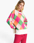 A woman wearing a colorful oversized knit sweater with a large diamond argyle pattern in pink, orange, green, and cream, paired with bright pink pants, against a plain white studio background.