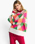 A woman modeling a colorful geometric knit sweater with pink, orange, green, and cream diamond patterns, paired with bright pink pants, against a plain white background.