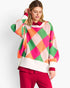 A woman wearing a colorful knit sweater with a large argyle pattern in pink, green, orange, and cream, paired with bright pink pants, against a plain light gray studio background.