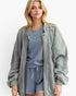 A blonde woman modeling a light sage green zip-up jacket over a matching dusty blue polka-dot lounge set against a plain white background.