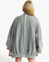 A woman viewed from behind wearing an oversized light gray jacket with a central vertical seam and patch pockets, against a plain white background.