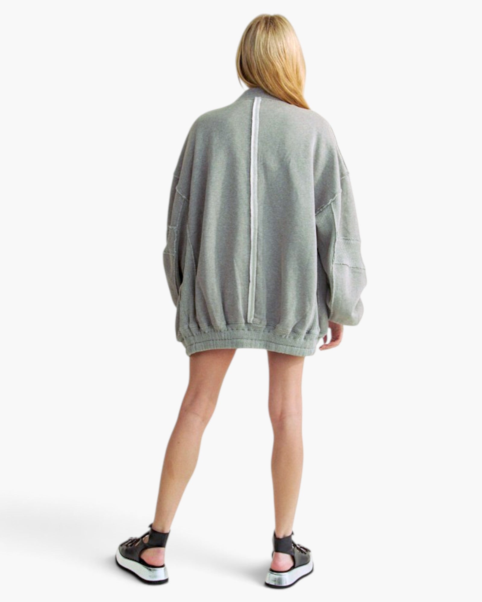 A woman shown from the back wearing an oversized light gray knit sweater with a prominent white vertical stripe down the center back, set against a plain light gray studio background.